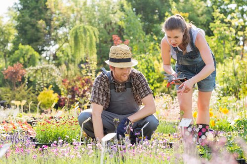 In a well-maintained garden, two individuals are engaged in gardening activities during daylight hours, with sunlight illuminating the vibrant natural surroundings. The scene features a variety of flowering plants with pink, purple, and yellow blooms in the foreground, bordered by lush green grass. Behind them, there are multiple trees, including a weeping willow with drooping branches, and dense foliage contributing to a secluded, peaceful outdoor space. The ground is a mix of flower beds with mulched soil and areas of neatly cut grass. One person, sitting on the grass, wears a straw hat, a checkered shirt, and gardening gloves, tending to small plants or flowers. The other individual, bending over, wears a sleeveless top and denim shorts, holding gardening tools and inspecting the plants. The background includes more flower beds, garden ornaments, and natural greenery, creating a vibrant and inviting environment that reflects active outdoor maintenance and gardening work typical of a private or community garden in Enfield. The scene visually aligns with gardening services provided by Gardening Services Enfield, emphasizing lawn and plant care in a peaceful residential outdoor setting.