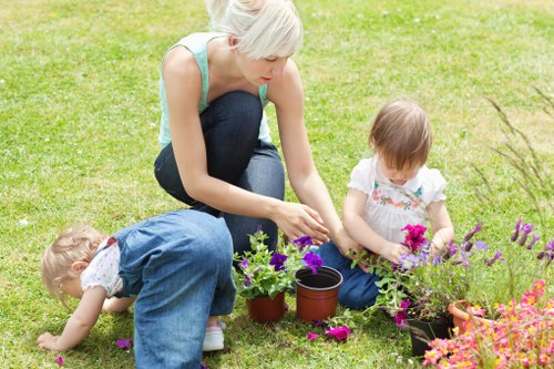 A woman with short blonde hair, wearing a light blue sleeveless top and dark jeans, is kneeling on a well-maintained lawn in a residential garden in Enfield. She is actively tending to various potted flowering plants, including purple petunias and pink blooms, which are arranged along the edge of the garden. Two young children, one with blond hair and dressed in denim overalls, are also engaged in gardening activities; the child on the left is bending down closely inspecting the grass or soil, while the other, with light brown hair and wearing a white dress with floral patterns, is sitting nearby with a small flower pot and a container of soil or seedlings. The background features a lush green hedge, a mix of small shrubs, and a section of paving or patio edging. The scene is illuminated by natural daylight, suggesting a clear, sunny day, consistent with typical outdoor maintenance and gardening work in Enfield. This image exemplifies family-friendly gardening activities, with an emphasis on plant care and outdoor enjoyment, aligned with services offered by Gardening Services Enfield for enhancing residential gardens and outdoor spaces.
