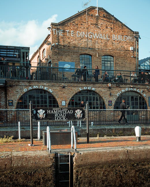 A historic brick building known as The Teddington Lock Building, with weathered red bricks and large arched windows, features signage indicating the Camden, Lock, and Duncalms areas. In front of the b