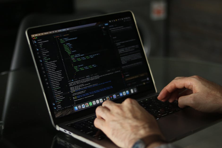 A close-up view of a person's hands typing on a sleek, silver laptop computer with a dark keyboard. The laptop screen displays lines of colorful code and programming text, indicating active software d