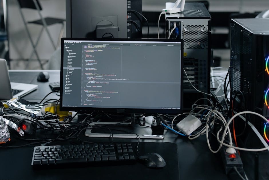 A cluttered desk featuring a computer monitor displaying lines of code, surrounded by various tangled cables, a keyboard, and a mouse. In the background, there are two desktop computer towers with vis